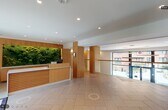 This contemporary two-floor lobby space combines refined design with welcoming functionality. The main entry greets visitors with large-format light tile flooring and a striking reception desk framed by warm wood paneling and a dramatic living moss wall installation, bringing a vibrant natural element to the interior. Recessed lighting throughout maintains a clean, bright ambiance.

The lobby extends into a spacious double-height atrium anchored by a sculptural circular chandelier suspended overhead. Wood-clad structural columns define the vertical reach of the space, while a glass-railed upper mezzanine level overlooks the ground floor below. Curated artwork, including a large-scale landscape painting, adds character to the open wall surfaces.

Seating areas are thoughtfully arranged throughout, featuring modern lounge chairs, sofas, and occasional tables atop geometric patterned area rugs. A secondary lounge area offers a built-in linear fireplace set within a floor-to-ceiling wood surround, creating an intimate retreat within the broader space.

Floor-to-ceiling glazed doors and expansive windows flood the lobby with natural light and frame views of an exterior landscaped courtyard complete with seating and greenery. A glass-balustrade staircase with brass accents provides elegant access between floors, reinforcing the space's polished, contemporary character throughout.
