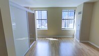 This single-floor unit offers a clean, functional layout with notable urban character. The main living area is finished with light-toned hardwood flooring that reflects natural light, contributing to an open and airy atmosphere. Two generously sized windows dominate one wall, framing elevated views of a dense city skyline — a standout feature that brings the energy of the surrounding urban environment directly into the space. The windows are fitted with horizontal blinds, allowing flexible light control throughout the day. The walls are finished in a warm neutral tone, complementing the light wood flooring and creating a cohesive, understated aesthetic. A mirrored sliding closet is positioned along one side of the room, serving both a practical storage function and visually expanding the sense of space by reflecting natural light and city views. White baseboards and trim provide clean contrast against the warmer wall tones. The overall ambiance is modern and minimal, well-suited for urban living. The elevated position of the unit, evidenced by the high-rise cityscape visible through the windows, adds a distinctive sense of place and connection to the surrounding metropolitan environment.