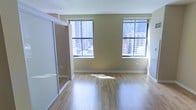 This single-floor unit offers a clean, functional layout with notable urban character. The main living area is finished with light-toned hardwood flooring that reflects natural light, contributing to an open and airy atmosphere. Two generously sized windows dominate one wall, framing elevated views of a dense city skyline — a standout feature that brings the energy of the surrounding urban environment directly into the space. The windows are fitted with horizontal blinds, allowing flexible light control throughout the day. The walls are finished in a warm neutral tone, complementing the light wood flooring and creating a cohesive, understated aesthetic. A mirrored sliding closet is positioned along one side of the room, serving both a practical storage function and visually expanding the sense of space by reflecting natural light and city views. White baseboards and trim provide clean contrast against the warmer wall tones. The overall ambiance is modern and minimal, well-suited for urban living. The elevated position of the unit, evidenced by the high-rise cityscape visible through the windows, adds a distinctive sense of place and connection to the surrounding metropolitan environment.