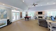 Located at 102 Village Dr in Mebane, NC, this single-story community clubhouse offers a well-appointed gathering space designed for both relaxation and productivity. The expansive great room serves as the heart of the facility, boasting soaring vaulted ceilings with recessed lighting and a ceiling fan, neutral carpet flooring, and a striking stacked-stone fireplace with a mounted flat-screen television above it — creating a warm and inviting focal point. Large windows throughout flood the space with abundant natural light and offer views of the surrounding greenery. A full-size billiards table anchors the central area, complemented by comfortable seating arrangements ideal for socializing. Adjacent to the main lounge, a wet bar features rich wood cabinetry, a mosaic tile backsplash, dark countertops, and bar-height seating — perfect for casual entertaining. French doors lead to an outdoor patio, seamlessly connecting the interior to the outdoors. A dedicated office nook, visible through glass-paned interior doors, provides a quiet workspace equipped with a desk and computer setup. The overall ambiance is warm and welcoming, blending functional amenities with comfortable, residential-style design throughout.
