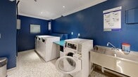 Located at 135 Allan St in Oakville, Ontario, this shared laundry facility is a thoughtfully designed, single-floor space that offers a functional and comfortable experience for its users. The room is finished with bold cobalt blue accent walls contrasted against crisp white ceilings adorned with crown moulding and recessed pot lighting, creating a clean and modern atmosphere.

The laundry area is well-equipped with multiple front-loading washing machines and top-loading dryers arranged along the walls, accompanied by teal-topped folding tables that provide ample workspace for sorting and folding laundry. A stainless steel utility sink is also available for hand-washing needs. Informational signage and a payment system are conveniently mounted on the walls for easy access.

A dedicated waiting area offers clear acrylic chairs and a small side table, providing a comfortable spot for residents while their laundry cycles complete. A wall clock keeps track of time, and a large, vivid ocean wave artwork adds a vibrant visual focal point to the space. A teal bookshelf with reading materials offers an added amenity for those waiting.

The terrazzo flooring throughout lends a durable and polished finish, tying the space together with a timeless aesthetic. The facility is also equipped with a security camera and fire safety systems, ensuring a safe and monitored environment.