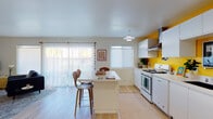 This cheerful single-floor residence blends retro-inspired design with modern finishes throughout its well-appointed layout. The kitchen serves as a visual centerpiece, featuring crisp white cabinetry, a stainless steel range hood, a gas range, and a bold yellow accent wall that adds striking warmth to the space. A white center island with mid-century style wood barstools provides a casual dining area, while sheer curtain-draped sliding glass doors flood the room with natural light and offer views of the outdoors. Light wood flooring flows seamlessly throughout the home, enhancing its bright, airy feel. The living room is thoughtfully designed with an eclectic, artistic character — an oversized arc floor lamp, bold wall art, floating shelves with greenery, and a yellow media console contribute to its lively personality. Both bedrooms are comfortably sized, bathed in natural light from large windows dressed with sheer curtains, and finished with warm wood floors and generous closet space. The two bathrooms are cleanly designed with white vanities, matte black fixtures, glass-enclosed showers, and classic white hexagonal tile flooring — offering a refined, contemporary feel that complements the home's vibrant overall aesthetic.