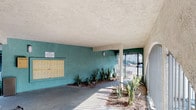 This multi-unit residential building showcases a practical, open-air design with stucco exterior walls finished in neutral cream and teal tones. The property is organized around a central courtyard-style layout, where paved walkways — featuring a mix of brick pavers and concrete — connect the various areas of the complex.

The covered ground-floor entry area serves as a functional common space, housing a bank of individual mailboxes mounted on a teal accent wall, providing convenient mail access for all residents. This shaded area also accommodates a dedicated bike storage section, where multiple bicycles can be secured under the protective overhang. Arched metal gates add a decorative architectural touch to the entry points, complementing the stucco columns that support the structure above.

Landscaped planting beds featuring drought-tolerant succulents and ornamental grasses are integrated throughout the common areas, adding natural texture and greenery. An exterior staircase leads to the upper level, where a walkway and metal railing balcony provide access to individual units. The open design of the common areas allows ample natural light to filter through, creating a bright and airy atmosphere throughout the shared spaces.