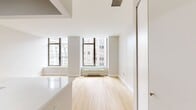 Located at 10 Hanover Place, Unit 21X in New York City, this 340-square-foot studio apartment offers a compact yet efficiently designed single-floor layout comprising an open-plan kitchen and living area along with one full bathroom. The living space is characterized by light-toned hardwood flooring throughout, crisp white walls, and large dark-framed windows that invite abundant natural light and frame elevated views of the surrounding Manhattan skyline. A baseboard heating unit runs beneath the windows, providing in-unit climate control. The kitchen is well-appointed with rich walnut-toned cabinetry, a white quartz countertop featuring an undermount stainless steel sink with a modern gooseneck faucet, subway tile backsplash, stainless steel appliances including a refrigerator, gas range, and microwave, and ample upper and lower cabinet storage. The bathroom is finished with large-format beige marble-style floor and wall tiles, a soaking tub with an overhead shower, a wood-grain vanity with a white countertop and undermount sink, a large mirror, and a toilet. The overall aesthetic is clean and contemporary, making efficient use of the available square footage in a prime Lower Manhattan location.