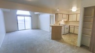 This single-floor space showcases an open-concept layout that seamlessly connects the living area and kitchen. The living area is finished with neutral-toned carpet and is bathed in natural light courtesy of a large arched window and a sliding glass door that spans a generous portion of one wall, offering expansive outdoor views and creating an airy atmosphere throughout.

The kitchen is positioned to the right and features warm wood-toned cabinetry with light countertops and a peninsula that provides a natural visual boundary between the two spaces without sacrificing the open feel. White appliances, including a gas range and dishwasher, are neatly integrated beneath the cabinetry. Tile flooring in the kitchen contrasts with the carpeted living space, clearly defining each functional zone.

A recessed transition area visible in the background hints at additional rooms beyond the main living space. Overhead lighting in both the kitchen and living area ensures the space remains well-lit. The overall ambiance is clean, neutral, and functional, offering a versatile layout suited to a variety of living arrangements.