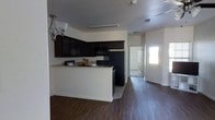 This single-story space showcases a well-designed open layout that seamlessly connects the kitchen and living areas. The kitchen is a standout feature, equipped with rich, dark espresso-toned cabinetry that extends to the upper wall, providing ample storage. The countertops display a light granite-look finish, offering a striking contrast against the dark cabinetry. Black appliances, including a refrigerator and built-in microwave, complement the kitchen's cohesive color scheme. A peninsula-style breakfast bar extends from the kitchen, creating a natural divide between the cooking and living spaces while offering additional seating and counter space. Dark hardwood-style flooring runs throughout, adding warmth and continuity to the interior. The walls are finished in a clean, neutral white, enhancing the brightness of the space. A decorative pendant light fixture with a classic design adds an elegant touch above the dining area. Toward the rear, a hallway leads to additional rooms, while a front entry door with a sidelight panel allows natural light to filter into the space, contributing to an inviting and functional ambiance.