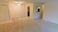 This single-floor residence offers a thoughtfully arranged layout that maximizes functionality and flow. The main living area is generously sized, featuring wall-to-wall carpeting in a neutral tone that lends a clean, understated look throughout the space. A ceiling fan with integrated lighting fixture serves as the central overhead feature, providing both comfort and illumination to the room.

The living area connects seamlessly to the kitchen, which is accessible through a dedicated entryway and further linked via a raised pass-through bar counter — an ideal feature for casual dining or entertaining. The kitchen is equipped with black appliances, including a gas range and a full-size refrigerator, alongside warm-toned cabinetry that provides ample storage. The pass-through window adds an open, airy quality to the kitchen-living relationship.

The walls throughout are finished in a light, neutral tone, contributing to a bright and welcoming atmosphere. The consistent use of carpet and coordinating wall colors creates a cohesive interior aesthetic. The layout is efficient, with clear sightlines between the living and kitchen areas, making the space feel open and well-connected. Overall, this residence offers a practical and comfortable environment well-suited for everyday living.