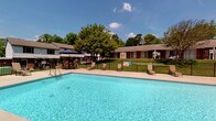 This outdoor pool area serves as a refreshing community amenity, designed exclusively for residents. The centerpiece is a generously sized swimming pool filled with clear, blue water, offering an inviting retreat during warm weather. Surrounding the pool is a spacious concrete deck that provides ample room for relaxation and movement.

Poolside seating includes several lounge chairs positioned to take advantage of the open sky and natural sunlight. A shaded outdoor dining area with a table, chairs, and a large umbrella offers a comfortable spot for gathering out of direct sun. Mature trees frame the area, adding greenery and a sense of privacy.

The pool area is fully enclosed by a black iron fence with a secure gate entrance, reinforcing its residents-only access. Signage at the entry points clearly designates the space as private. The brick exterior of the adjacent building adds a classic architectural character to the setting.

Well-maintained green lawn areas border the pool enclosure, contributing to the overall pleasant outdoor atmosphere. The surrounding community buildings, with their brick facades and low-profile rooflines, create a cohesive neighborhood backdrop. The combination of functional amenities, open-air layout, and lush surroundings makes this pool area a welcoming outdoor destination for residents.