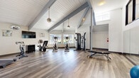 Located at 4255 NE 125th Place in Portland, Oregon, this two-level fitness facility offers a well-equipped workout environment spread across distinctly designed floors. The upper level is immediately distinguished by its striking vaulted ceiling with exposed gray beams and white shiplap paneling, creating a bright, airy atmosphere. Natural light streams in through multiple windows, some offering views of an outdoor pool area. Hardwood-style flooring runs throughout this level, which houses an array of cardio equipment including treadmills, ellipticals, a rowing machine, and a spin bike, alongside a cable resistance machine and adjustable bench. Pendant light fixtures and track lighting complement the natural light, while a wall-mounted television adds convenience. The lower level contrasts with a sleek, modern aesthetic featuring recessed lighting, carpeted flooring, and a clean white palette with dark trim accents. This floor focuses on strength training, with selectorized weight machines, a dumbbell rack, and additional cable equipment. Large mirrors amplify the sense of space, and a wall-mounted TV and mini-split HVAC unit enhance comfort. Stability balls and medicine balls are also available, supporting diverse workout routines throughout the facility.