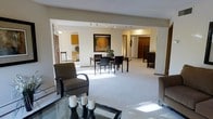 This single-floor space offers an open and airy layout that seamlessly connects the living, dining, and kitchen areas. The living area is anchored by plush wall-to-wall carpeting and features comfortable seating alongside a glass-topped coffee table, creating an inviting space for relaxation. A dedicated dining area accommodates a dark wood table and chairs, accented by a striking framed artwork depicting warm autumn foliage, adding a pop of color to the neutral surroundings. Track lighting runs along the ceiling, providing adjustable illumination throughout the open-plan space. The kitchen area, visible from the main living zone, features warm wood cabinetry and connects naturally to the rest of the floor plan. A hallway leads to additional rooms, hinting at a thoughtfully organized layout. The overall ambiance is calm and welcoming, characterized by a consistent neutral palette of whites and beiges that enhances the sense of openness. Natural light filters into the space, brightening the carpeted floors and contributing to the light, comfortable feel that defines this well-appointed single-level home.