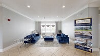 Located at 6334 Clock Tower Dr in Walbridge, Ohio, this community clubhouse entrance offers a polished and functional single-floor layout designed for both comfort and practicality. The space is finished with light neutral-toned walls and large-format ceramic tile flooring throughout, creating a clean, cohesive aesthetic. Crown molding accents the ceilings, adding a refined architectural detail that elevates the overall ambiance.

The main lounge area is generously proportioned, centered around a pair of tufted navy blue sofas and a matching oversized ottoman atop a vintage-style area rug. Accent chairs and side tables round out the seating arrangement, providing an inviting space for residents to gather. Recessed lighting illuminates the room evenly, while a bank of windows dressed with draped curtains allows natural light to filter in.

A dedicated workspace area accommodates a computer station and multifunction printer, offering practical amenities for residents. A secondary seating nook features dark leather furniture, providing a quieter setting for relaxed conversations or informal meetings.

The entrance foyer is anchored by a prominent white column and features glass double doors marked with a branded Queens Lake Communities welcome mat. The layout connects seamlessly between functional zones, making this an accessible and well-organized community hub.