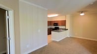 This single-floor space showcases an open-concept design that seamlessly connects the living area to the kitchen. The living area is finished with wall-to-wall neutral-toned carpet, creating a warm and comfortable atmosphere, while crown molding along the ceiling adds a refined architectural touch throughout.

The kitchen is a functional and well-appointed space, featuring rich medium-toned wood cabinetry that provides ample storage. Dark granite-style countertops offer a striking contrast against the light walls and white lower cabinetry of the peninsula. The kitchen is equipped with black appliances, including a range and an over-the-range microwave. A peninsula with a built-in sink serves as both a practical workspace and a natural divider between the kitchen and the adjoining living area, making it ideal for casual dining or entertaining.

The overall color palette of the space is light and neutral, with cream-toned walls that enhance the sense of openness. Overhead flush lighting in the kitchen ensures the workspace is well-illuminated. The layout is practical and efficient, with a smooth transition between the carpeted living zone and the hard-floored kitchen area.