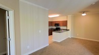 This single-floor space showcases an open-concept design that seamlessly connects the living area to the kitchen. The living area is finished with wall-to-wall neutral-toned carpet, creating a warm and comfortable atmosphere, while crown molding along the ceiling adds a refined architectural touch throughout.

The kitchen is a functional and well-appointed space, featuring rich medium-toned wood cabinetry that provides ample storage. Dark granite-style countertops offer a striking contrast against the light walls and white lower cabinetry of the peninsula. The kitchen is equipped with black appliances, including a range and an over-the-range microwave. A peninsula with a built-in sink serves as both a practical workspace and a natural divider between the kitchen and the adjoining living area, making it ideal for casual dining or entertaining.

The overall color palette of the space is light and neutral, with cream-toned walls that enhance the sense of openness. Overhead flush lighting in the kitchen ensures the workspace is well-illuminated. The layout is practical and efficient, with a smooth transition between the carpeted living zone and the hard-floored kitchen area.
