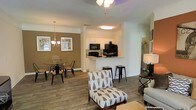 This single-floor residence offers an open-concept layout that seamlessly connects the living, dining, and kitchen areas into one cohesive and functional space. Rich hardwood-style flooring runs throughout, anchoring the warm, welcoming ambiance of the interior. The living area is generously sized, providing ample room for comfortable furnishings and relaxation, while a ceiling fan adds both style and practicality overhead. Adjacent to the living space, the dining area features an elegant chandelier and is set against a warm earth-toned accent wall, creating a cozy atmosphere for meals and gatherings. The kitchen is thoughtfully positioned at the heart of the home, featuring white cabinetry, black appliances, and a raised breakfast bar with barstool seating — ideal for casual dining or entertaining. Crown molding and decorative columns add a refined architectural touch, elevating the overall design of the space. Natural light filters into the dining area through a window, brightening the interior. The layout is both practical and aesthetically cohesive, with consistent finishes and warm tones that contribute to an inviting, comfortable living environment throughout the home.