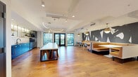 Located at 2534 W Colfax Ave in Denver, CO, this thoughtfully designed single-floor amenity space offers a generous and versatile layout across its open floor plan. Warm hardwood flooring runs throughout, unifying the distinct functional zones within the space.

The full-service kitchen area is a standout feature, outfitted with deep navy cabinetry accented by brass hardware and open brass-framed shelving with under-cabinet lighting. A large terrazzo communal dining table anchors the kitchen zone, surrounded by modern seating and illuminated by elegant curved pendant lighting. Stainless steel appliances, including a refrigerator and built-in ovens, complete the well-appointed culinary setup. Glass doors at the far end of the kitchen area open to an outdoor space, flooding the interior with natural light and city views.

Adjacent to the kitchen, a dining and social lounge area features banquette seating in warm caramel tones against a dramatic abstract mural wall, with pendant lighting adding a sophisticated ambiance. A separate, more intimate lounge zone provides plush seating arranged around a sculptural stone coffee table, anchored by a tile-framed fireplace and large-screen display.

A flexible open room with a striking floor-to-ceiling navy upholstered panel wall with gold trim detailing offers additional event or activity space. Curated artwork is displayed throughout, reinforcing the space's polished, design-forward character.