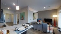 This single-floor space showcases a thoughtfully designed open-concept layout that integrates the dining and living areas into one cohesive environment. The dining area is anchored by a sleek granite or stone-topped counter, set with place settings beneath a contemporary pendant light that adds a warm, focused glow to the space. The light gray walls throughout create a neutral, modern backdrop that enhances the brightness of the interior.

The living area flows naturally from the dining space, featuring wood-look flooring that runs continuously through both zones, adding warmth and visual continuity. A gray sectional sofa is paired with a glass and chrome coffee table, lending a clean, contemporary feel. Colorful wall art adds personality and a pop of vibrancy against the neutral tones. Recessed lighting complements the pendant fixture, ensuring the space is well-lit throughout.

A hallway visible from the main living area leads to additional rooms, including what appears to be a bathroom, suggesting a well-organized floor plan. The open layout and cohesive design choices make this space feel inviting, functional, and well-suited for both everyday living and casual entertaining.