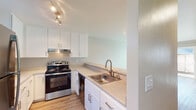 This single-floor unit offers a bright and functional layout highlighted by warm hardwood-style flooring that flows seamlessly throughout the living spaces. The open-concept living area is generously sized, featuring neutral-toned walls, clean white baseboards, and track lighting that provides ample illumination. A pass-through counter connects the living area to the kitchen, enhancing the sense of openness and making the space feel cohesive.

The kitchen is efficiently designed in an L-shaped configuration and features crisp white shaker-style cabinetry with brushed nickel hardware, light-toned countertops, and a stainless steel single-basin sink with a modern gooseneck faucet. Appliances include a stainless steel electric range with an overhead exhaust hood and a stainless steel refrigerator, providing a well-equipped cooking environment. The kitchen also benefits from track lighting and hardwood-style flooring consistent with the rest of the unit.

The bedroom is a comfortable, well-proportioned room with neutral warm-gray walls and a window that allows natural light and views of the surrounding greenery. An in-unit stacked washer and dryer is conveniently located in a dedicated hallway area, adding significant practical value. Overall, the unit presents a clean, move-in-ready aesthetic with a cohesive and modern design sensibility throughout.