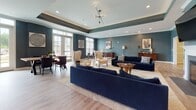 Located at 3120 Crossroads Station Blvd in Fredericksburg, Virginia, this single-floor community clubhouse offers a refined and welcoming environment designed for both relaxation and social gathering. The space is defined by rich teal-blue accent walls, tray ceilings with recessed lighting, and light wood-look flooring throughout, creating a cohesive and sophisticated aesthetic.

The main lounge area serves as the centerpiece of the space, featuring a classic white fireplace flanked by a mounted flat-screen TV, plush navy blue sofas, stylish upholstered armchairs, and gold-accented side tables. Large windows and French doors allow generous natural light to fill the room, while a sculptural spur chandelier adds a contemporary focal point overhead. A secondary seating area provides additional intimate groupings of chairs and sofas, ideal for casual conversation or quiet relaxation.

The kitchen is exceptionally well-appointed, boasting an extended granite countertop island with bar seating, dark cabinetry with brushed nickel hardware, stainless steel appliances, glass-front upper cabinets with open display shelving, and elegant pendant lighting. The kitchen flows seamlessly into the social areas, making it well-suited for catered events or community gatherings. Throughout the space, thoughtful décor including framed artwork and live greenery enhances the warm, upscale ambiance.