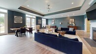 Located at 3120 Crossroads Station Blvd in Fredericksburg, Virginia, this single-floor community clubhouse offers a refined and welcoming environment designed for both relaxation and social gathering. The space is defined by rich teal-blue accent walls, tray ceilings with recessed lighting, and light wood-look flooring throughout, creating a cohesive and sophisticated aesthetic.

The main lounge area serves as the centerpiece of the space, featuring a classic white fireplace flanked by a mounted flat-screen TV, plush navy blue sofas, stylish upholstered armchairs, and gold-accented side tables. Large windows and French doors allow generous natural light to fill the room, while a sculptural spur chandelier adds a contemporary focal point overhead. A secondary seating area provides additional intimate groupings of chairs and sofas, ideal for casual conversation or quiet relaxation.

The kitchen is exceptionally well-appointed, boasting an extended granite countertop island with bar seating, dark cabinetry with brushed nickel hardware, stainless steel appliances, glass-front upper cabinets with open display shelving, and elegant pendant lighting. The kitchen flows seamlessly into the social areas, making it well-suited for catered events or community gatherings. Throughout the space, thoughtful décor including framed artwork and live greenery enhances the warm, upscale ambiance.
