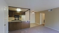 This single-floor residence offers an open and functional layout that connects the kitchen and living areas in a cohesive design. The kitchen is equipped with dark wood cabinetry, a built-in microwave, an electric range, and a white refrigerator. A breakfast bar with a dark wood fascia extends from the kitchen, providing a casual dining or workspace option and naturally defining the boundary between the kitchen and the living area. A decorative twisted column detail adds a distinctive architectural touch to this partition. The living area is finished with wall-to-wall carpet in a neutral tone, creating a warm and comfortable atmosphere. A ceiling fan with a light fixture is centrally positioned overhead, offering both air circulation and ambient lighting. Neutral-toned walls and white baseboards keep the space feeling clean and cohesive throughout. A hallway visible from the main living area leads to additional rooms, suggesting a practical and well-organized floor plan. The textured ceiling is consistent throughout the space, and overall the residence presents a straightforward, move-in-ready layout suited for everyday living.