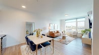 This single-floor unit showcases a contemporary open-concept layout with light hardwood-style flooring flowing throughout. The combined living and dining area is bright and welcoming, anchored by a large window that frames sweeping city views and floods the space with natural light. A seating nook positioned beside the oversized window offers an ideal spot to take in the urban panorama.

The kitchen is efficiently designed with warm wood cabinetry extending to the ceiling, stainless steel appliances including a refrigerator, range, microwave, and dishwasher, and a decorative white tile backsplash. Light countertops and generous storage make the kitchen both functional and visually clean.

The bedroom is a calm retreat, featuring a large window with outdoor views, wall-mounted reading sconces, and direct access to a well-organized walk-in closet with built-in shelving and hanging rods. The full bathroom features wood-toned cabinetry, light wood-look flooring, and a combined tub and shower. Crisp white walls and recessed lighting contribute to the unit's clean, modern aesthetic throughout.