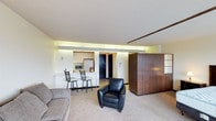 This single-floor space offers an open-concept design that seamlessly integrates the living area with a compact kitchenette, creating a functional and cohesive layout. The living area is carpeted and furnished with a plush sofa and a sleek black leather armchair, providing a comfortable seating arrangement. A prominent dark wood partition or wardrobe unit serves as a stylish room divider, adding warmth and visual interest to the space. The kitchenette is efficiently organized with light wood cabinetry, a refrigerator, a microwave, and a breakfast bar with two bar-height chairs, making it ideal for casual dining. Recessed cove lighting runs along the ceiling perimeter, casting a soft, ambient glow that enhances the modern aesthetic. The entryway features a dark front door, and the overall color palette is neutral, with cream-toned walls and light ceilings contributing to a bright and inviting atmosphere. The textured ceiling adds a subtle architectural detail throughout. The layout maximizes the available footprint, ensuring every area is purposeful and well-utilized, making this a practical and contemporary single-floor space.