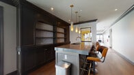 Located at 936 N High St in Columbus, Ohio's vibrant Short North neighborhood, this single-floor space makes a strong design statement through its carefully curated interior. The most prominent feature is a striking bar and kitchen area defined by deep, dark-toned custom cabinetry with arched open shelving backed by marble-look stone panels. A substantial central island with a sleek stone countertop and brass accent trim anchors the space, lined with warm cognac leather barstools on gold-toned bases. Hanging above are elegant cylindrical brass pendant lights that cast a warm, refined glow, complementing the rich hardwood flooring throughout. A built-in sink and ample counter space make the area as functional as it is visually impressive. The open sightlines extend toward an adjacent lounge area, hinting at the broader layout beyond. Additional rooms throughout the space feature windows with blinds that filter in natural light, contributing to the overall atmosphere. The combination of dark finishes, metallic accents, and high-end materials throughout creates a sophisticated, upscale ambiance well-suited for both entertaining and everyday use in one of Columbus's most dynamic neighborhoods.