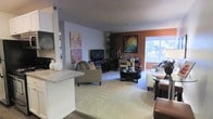 This single-floor residence offers an open-concept layout that connects the living, dining, and kitchen areas into one cohesive and functional space. The living area is anchored by plush carpeting and features a comfortable seating arrangement centered around a stylish round coffee table, with a media console and flat-screen television set against the wall. Decorative canvas artwork in soft floral tones adds a personal touch to the light gray walls, while a bold terracotta accent wall provides a warm contrast and serves as a visual focal point. A large window bathes the space in abundant natural light, offering views of the greenery outside and creating an airy, open atmosphere. A tall indoor plant adds a refreshing organic element to the corner of the room. Adjacent to the living area, a dining space accommodates a small table and chairs, ideal for casual meals or entertaining. The kitchen is partially visible beyond a breakfast bar with a light stone countertop, hinting at a practical and accessible layout. The combination of warm accent colors, soft textures, and natural light gives the entire space a comfortable and welcoming ambiance throughout.