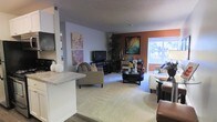This single-floor residence offers an open-concept layout that connects the living, dining, and kitchen areas into one cohesive and functional space. The living area is anchored by plush carpeting and features a comfortable seating arrangement centered around a stylish round coffee table, with a media console and flat-screen television set against the wall. Decorative canvas artwork in soft floral tones adds a personal touch to the light gray walls, while a bold terracotta accent wall provides a warm contrast and serves as a visual focal point. A large window bathes the space in abundant natural light, offering views of the greenery outside and creating an airy, open atmosphere. A tall indoor plant adds a refreshing organic element to the corner of the room. Adjacent to the living area, a dining space accommodates a small table and chairs, ideal for casual meals or entertaining. The kitchen is partially visible beyond a breakfast bar with a light stone countertop, hinting at a practical and accessible layout. The combination of warm accent colors, soft textures, and natural light gives the entire space a comfortable and welcoming ambiance throughout.