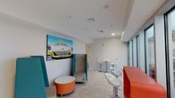 The Lounge is a stylish and energetic communal space designed to encourage relaxation and social interaction. The interior is finished with clean white walls and light-toned tile flooring throughout, creating a bright and open atmosphere. A striking pop-art canvas depicting a classic automobile serves as a bold focal point, injecting vibrant color and personality into the space.

Seating is thoughtfully varied to accommodate different preferences and group sizes. High-back teal lounge chairs paired with a round orange ottoman offer a cozy, semi-private nook, while modern white bar-height tables and swivel stools provide a more casual, upright option for conversation or light work. A bold red bar unit with matching bar stools adds another dynamic seating area near the expansive glazed wall. A wall-mounted flat-screen television is also present, adding to the lounge's entertainment appeal.

One of the most compelling features of the space is the full wall of floor-to-ceiling glass sliding doors, which floods the interior with natural light and provides direct access to a generous outdoor terrace. The terrace extends the usable space considerably and offers open sky views, making it an ideal setting for gatherings or quiet moments outdoors. Recessed ceiling lighting and integrated speakers enhance the ambiance throughout. Overall, The Lounge strikes a balance between lively design and functional comfort.