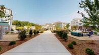 Located at 10315 Monrovia Dr in Matthews, NC, this residential community showcases a well-planned outdoor environment designed for both functionality and aesthetic appeal. The grounds are anchored by a broad, central concrete walkway that stretches through the heart of the community, flanked by neatly arranged young trees, manicured shrubs, and fresh mulched beds that lend a clean, welcoming character to the exterior.

The multi-story residential buildings surrounding the courtyard feature a mix of architectural styles, incorporating elements such as lap siding in neutral and teal tones, brick accents, gabled rooflines, and balconies. These design choices create a cohesive yet visually engaging streetscape. A covered porte-cochère or breezeway area is prominently featured, supported by tall white columns and finished with arched entryways that lead into glazed interior spaces, adding an elegant transitional element between the outdoors and interior.

Outdoor amenity areas are woven throughout the grounds, including seating nooks and what appears to be a hammock station nestled among the trees, encouraging residents to enjoy the open-air environment. Wide concrete patios run along the building facades, providing accessible pathways and gathering spaces. The overall ambiance is bright, open, and community-oriented, with abundant natural light and clear skies enhancing the inviting atmosphere of this Matthews neighborhood.