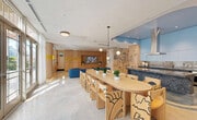 Located at 2250 NW 1st Ave in Miami's creative Wynwood district, this single-floor space is a dynamic blend of art, functionality, and modern design. The open-plan layout connects a fully equipped kitchen, a generous dining area, and a comfortable lounge, creating a seamlessly flowing communal environment.

The kitchen features a striking dark granite island and countertops, sleek light-blue cabinetry, and a prominent stainless steel range hood, all set beneath recessed lighting and a soft blue ceiling. Adjacent to it, a large oval communal dining table surrounded by artisan light wood chairs — each uniquely decorated with bold black illustrated motifs — serves as a natural gathering focal point, illuminated by sculptural pendant lights.

The lounge area is anchored by a deep blue sectional sofa, a round wood coffee table, and a colorful area rug, set against a wall of warm natural wood paneling with perforated yellow accents and a mounted flat-screen TV. Floor-to-ceiling sheer curtains filter abundant natural light from large windows, adding an airy ambiance.

Throughout the space, expressive hand-drawn murals animate the walls with a playful, urban energy reflective of Wynwood's artistic character. Polished terrazzo floors tie the entire space together, while a cozy nook with white armchairs and vivid accent rugs provides an intimate retreat within this vibrant, community-centered space.