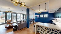 Located at 555 Pacific Ave in Santa Cruz, CA, this single-floor communal amenity space offers a thoughtfully designed open-plan layout that seamlessly integrates a living area, dining space, and kitchenette. The interior is defined by striking cobalt blue accent walls and columns that contrast boldly against neutral white tones, creating a vibrant and modern aesthetic throughout.

The living area is anchored by a full-size pool table atop a distinctive herringbone-patterned hardwood floor, complemented by a plush sectional sofa, leather lounge chairs, and a wall-mounted flat-screen TV. Contemporary pendant and sputnik-style chandeliers provide warm, stylish illumination. Large floor-to-ceiling glass French doors flood the space with natural light and open directly onto an outdoor courtyard patio.

The kitchenette features sleek dark cabinetry with black hardware, a white countertop peninsula with bar seating, stainless steel appliances including a refrigerator and microwave, and an under-counter sink — ideal for hosting and gatherings. A built-in wall-length wooden counter with bar stools beneath dual windows serves as a casual dining and workspace area, offering views of the surrounding greenery.

Together, these zones create a versatile, social environment with a cohesive modern design perfectly suited for community use.