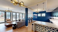 Located at 555 Pacific Ave in Santa Cruz, CA, this single-floor communal amenity space offers a thoughtfully designed open-plan layout that seamlessly integrates a living area, dining space, and kitchenette. The interior is defined by striking cobalt blue accent walls and columns that contrast boldly against neutral white tones, creating a vibrant and modern aesthetic throughout.

The living area is anchored by a full-size pool table atop a distinctive herringbone-patterned hardwood floor, complemented by a plush sectional sofa, leather lounge chairs, and a wall-mounted flat-screen TV. Contemporary pendant and sputnik-style chandeliers provide warm, stylish illumination. Large floor-to-ceiling glass French doors flood the space with natural light and open directly onto an outdoor courtyard patio.

The kitchenette features sleek dark cabinetry with black hardware, a white countertop peninsula with bar seating, stainless steel appliances including a refrigerator and microwave, and an under-counter sink — ideal for hosting and gatherings. A built-in wall-length wooden counter with bar stools beneath dual windows serves as a casual dining and workspace area, offering views of the surrounding greenery.

Together, these zones create a versatile, social environment with a cohesive modern design perfectly suited for community use.