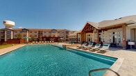 Located at 255 Williamsburg Pkwy in Fate, Texas, this expansive outdoor amenity space serves as the centerpiece of a modern residential community. Spanning a generous footprint, the area is anchored by a large, resort-style swimming pool with crystal-clear blue water, framed by a broad stone-paved sun deck lined with chaise lounge chairs and shaded umbrella stations — ideal for relaxing under the Texas sun. Adjacent to the pool, a handsome pergola structure provides a shaded gathering area, complemented by outdoor seating and a grilling station for social entertaining. The clubhouse building, constructed with warm stone and wood accents beneath a pitched roof with dramatic peaked gables, features floor-to-ceiling windows that flood the interior with natural light while offering views of the pool area. A second pool area on the property expands the aquatic offerings, surrounded by additional lounge seating, turfed lawn areas, and a perimeter fence with clearly posted safety signage. Throughout the grounds, manicured green lawns and young trees add natural texture and soften the built environment. The multi-story residential buildings surrounding the amenity areas are finished in a contemporary mix of siding, brick, and board-and-batten, with private balconies overlooking the landscaped courtyards. The overall ambiance is that of a well-appointed, community-focused outdoor retreat.