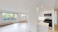Unit 308 at 2400 New St in Burlington, Ontario presents a thoughtfully renovated single-floor residence finished in a clean, contemporary palette. Light wood-look flooring runs consistently throughout, complementing the crisp white walls and trim that give the space an airy, cohesive feel.

The open-concept living area is a standout feature, anchored by large windows and a glass-paned door that opens to a private balcony. Treetop views of mature greenery are visible from multiple vantage points, bringing the outdoors in and enhancing the sense of openness. Baseboard heating runs beneath the windows, ensuring practical comfort year-round.

The kitchen is modern and well-appointed, featuring high-gloss white cabinetry extending to the ceiling, light quartz countertops, a double undermount sink, and stainless steel appliances including a refrigerator, microwave, and range. Recessed pot lighting illuminates the workspace effectively, while a peninsula provides additional counter space and separation from the living area.

The bedrooms are quietly positioned, each finished with the same light flooring and neutral tones. Both rooms feature mirrored sliding closet doors with built-in shelving and wire organizers, maximizing storage. Windows in the bedrooms frame leafy outdoor views, contributing to a calm and restful atmosphere throughout the unit.