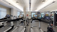 This single-level fitness center offers a comprehensive workout environment designed to accommodate a wide range of training needs. The space is organized into distinct functional zones, providing ample room for movement between equipment. A suspended tile ceiling with linear fluorescent lighting ensures consistent illumination throughout, while multiple windows fitted with horizontal blinds introduce natural light and views of outdoor greenery.

The cardio zone is prominently featured, housing a row of Life Fitness treadmills equipped with individual touchscreen monitors, alongside elliptical cross-trainers and a Peloton stationary bike. A rowing machine adds further cardiovascular variety. Strategically placed mirrors along the walls visually expand the space and allow users to monitor their form during workouts.

The strength training area includes a multi-station cable machine, a rack system, and an organized dumbbell rack spanning a comprehensive weight range. Medicine balls, stability balls, battle ropes, and weighted slam balls are also available for functional and dynamic training. Framed artwork adorns the walls, contributing a polished aesthetic to the otherwise utilitarian environment. The rubber-floored surfaces throughout provide safety and durability, while a wall-mounted television keeps users informed and entertained during sessions.