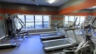 This dedicated fitness center is designed to provide a comprehensive workout environment within a single, well-organized space. The room is finished with bold, energizing décor — bold orange and gray horizontal stripes run along the upper walls, creating a dynamic and motivating atmosphere that complements the active purpose of the space. The flooring is a durable, dark rubberized surface well-suited for exercise use. A row of treadmills is positioned along the front wall, facing a bank of large windows that allow generous natural light to fill the room and offer views of the surrounding greenery, creating an inviting setting for cardio workouts. Additional strength and resistance equipment, including a cable machine, rounds out the selection of available fitness options. A full-length mirror spans one side wall, both enhancing the sense of space and allowing users to monitor their form during exercise. A ceiling fan overhead aids in air circulation, contributing to comfort during workouts. The layout is thoughtfully arranged to maximize floor space and equipment accessibility, making the fitness center a practical and well-appointed amenity for daily use.