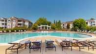 Located at 1 Madison Park Dr in Madison, Alabama, this expansive outdoor amenity space offers a resort-style experience across a generously sized single-level footprint. The centerpiece is a freeform swimming pool with a distinctive dual-lobe shape, providing ample surface area for leisure and recreation. The pool deck is finished in warm-toned concrete and is lined with modern chaise lounges and side tables, offering plenty of room for sunbathing and relaxation beneath the open Alabama sky.

A striking white pergola serves as a focal architectural element, framing a shaded seating area with paver flooring and additional lounge seating. Surrounding the pool area, mature trees, ornamental grasses, layered shrubbery, and stone retaining walls create a lush, well-maintained landscape that adds privacy and natural beauty to the environment.

The clubhouse entrance is equally impressive, featuring a traditional exterior with stone column bases, white-framed floor-to-ceiling glass panels, a covered porch with classical columns, and broad concrete steps that provide a welcoming and refined arrival experience. Carefully curated foundation plantings with trimmed hedges and cypress trees complete the polished aesthetic. The overall ambiance is that of a well-appointed, resort-caliber outdoor retreat.