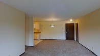 This single-floor residence offers a straightforward and functional layout designed for comfortable everyday living. The main living area is finished with wall-to-wall carpeting in a neutral tone, creating a warm and understated atmosphere throughout the space. Smooth, light-colored walls contribute to a clean and open feel, maximizing the sense of space within the unit.

The living area flows seamlessly into a compact kitchen, which is partially open to the main room. The kitchen features white cabinetry providing ample storage, a countertop with a utility sink, and is illuminated by a stylish pendant light fixture that hangs from the ceiling — adding a subtle decorative touch to an otherwise practical space. A flush-mounted ceiling light near the entryway provides additional illumination.

The entryway is marked by a solid dark-toned door, offering a strong visual contrast against the lighter walls and lending a sense of security and privacy. Adjacent to the front door, a closet with a sliding door provides convenient storage right at the entry point of the unit.

Overall, the space presents a clean, move-in-ready environment with a neutral palette and functional design, well-suited for those seeking a practical and low-maintenance living space on a single level.