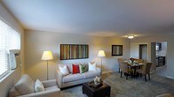 This single-story space showcases an open-plan layout that seamlessly connects the living and dining areas, creating a cohesive and functional environment. The living area is anchored by a light-toned sofa adorned with a mix of decorative pillows in white, red, and green, complemented by a dark upholstered ottoman that serves as a central coffee table. Twin floor lamps flank the seating arrangement, providing warm ambient lighting. A bold, colorful striped artwork commands attention on the main wall, adding a vibrant focal point to the otherwise neutral color palette of soft grays and warm whites. The smooth, light-colored ceiling enhances the sense of openness throughout the space. Transitioning naturally from the living area, the dining space accommodates a table surrounded by upholstered chairs, with a mirror mounted on the wall that reflects light and adds visual depth. A fresh floral arrangement on the dining table introduces a touch of color. The flooring transitions from carpet in the living zone to a harder surface in the dining area, subtly delineating the two spaces while maintaining an overall sense of continuity and flow.