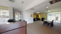 The Parkside Apartments leasing office is a well-organized, single-floor community facility designed to welcome and serve residents and prospective tenants alike. The space is anchored by a bright, glass-enclosed entrance lobby featuring floor-to-ceiling windows and double glass doors that flood the area with natural light and offer clear views of the surrounding streetscape. A polished terrazzo floor and a dedicated reception desk positioned near the entrance provide a professional first impression, making it easy for visitors to be greeted promptly upon arrival.

Adjacent to the reception area is a carpeted meeting zone furnished with a wooden table and dark chairs, ideal for leasing consultations or small group gatherings. The neutral gray and white tones of this area create a calm, professional atmosphere.

Beyond the front office, the space transitions into a generously sized resident lounge finished in warm sage green. This comfortable area is furnished with dark leather sofas, accent chairs, and side tables with lamps, creating an inviting retreat. The lounge is well-equipped with a wall-mounted flat-screen television, a water cooler, a full-size refrigerator, and a microwave — offering practical amenities for resident use. Artwork on the walls and thoughtful lighting add character and warmth throughout, making this leasing office both functional and welcoming.