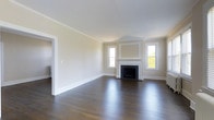 This single-floor space showcases a thoughtfully designed living area that combines classic architectural details with a clean, understated aesthetic. The room is anchored by a traditional fireplace framed with a crisp white surround and a decorative mantel, adding a focal point of timeless character. Flanking the fireplace are two windows that allow natural light to fill the room, highlighting the rich, dark hardwood floors that run throughout the space. The floors are polished to a reflective finish, contributing to a sense of depth and warmth. Neutral, warm beige walls are accented by well-defined crown molding and substantial white baseboards, reinforcing the traditional architectural style. The ceiling height adds to the overall sense of openness, making the room feel spacious and airy. A doorway visible on the left wall suggests additional connected spaces, offering functional flow throughout the layout. The overall ambiance is calm and refined, with a neutral palette that provides a versatile backdrop suitable for a variety of interior styles.