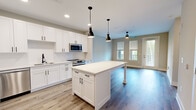 This thoughtfully designed one-bedroom, one-bathroom residence in Jacksonville, FL (32256) offers an efficient and functional single-floor layout with a clean, modern aesthetic throughout. The open-concept kitchen is a standout feature, showcasing white shaker cabinetry, a white quartz countertop island with pendant lighting, a subway tile backsplash, and a full suite of stainless steel appliances including a refrigerator, range, microwave, and dishwasher. Light wood-tone flooring runs through the main living areas, complementing the neutral color palette and creating a bright, cohesive feel. Large windows with blinds allow natural light to fill the living area, which also features a ceiling fan and access to an exterior door. The bedroom offers plush carpeting, a ceiling fan, and windows with outdoor views of surrounding greenery, providing a calm and comfortable retreat. The bathroom is well-appointed with gray shaker-style cabinetry, a white quartz vanity top, a large framed mirror, and a glass-enclosed shower. Directly accessible from the bathroom, the walk-in closet features wire shelving and carpeted flooring, offering generous storage. This residence combines contemporary finishes with a practical layout suited for comfortable everyday living.