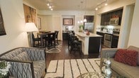 This single-floor open-concept space seamlessly integrates the living, dining, and kitchen areas into one cohesive and well-appointed layout. Dark hardwood floors run throughout, providing a rich, grounding contrast to the light-toned walls and cabinetry. The kitchen serves as a central hub, anchored by a large island with a white quartz countertop and dark cabinetry below, offering seating for casual dining. Stainless steel appliances, including a French door refrigerator and built-in microwave, add a modern, professional touch. Pendant lights hang above the island, while track lighting illuminates the space from above. Adjacent to the kitchen, the dining area features a dark wood high-top table with matching bar-height chairs, positioned atop a patterned area rug. Framed artwork on the walls adds personality. The living area, defined by a plush striped area rug, includes patterned upholstered seating and a glass-top coffee table, creating an inviting and stylish environment. The overall ambiance is contemporary and well-balanced, with a cohesive dark-and-light color palette throughout.