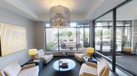 This elegant single-floor space presents a polished and welcoming lobby lounge designed with a refined contemporary aesthetic. The centerpiece of the room is a striking large-scale drum chandelier with crystal accents, suspended from a recessed ceiling that adds a sense of height and grandeur to the space. The chandelier casts a warm, inviting glow that complements the natural light flooding in through expansive floor-to-ceiling windows along the exterior wall. These large windows frame views of a landscaped outdoor area and surrounding streetscape, creating a bright and airy atmosphere throughout. The seating arrangement is thoughtfully curated, featuring plush cream-toned upholstered armchairs and sofas grouped around a sleek circular glass coffee table, providing a comfortable and stylish gathering area. Accent table lamps with vibrant yellow shades punctuate the neutral color palette, adding subtle pops of warmth and color. Dark-finished side tables and low-profile furniture contribute to the modern, sophisticated tone of the space. The dark carpet grounds the room and contrasts elegantly with the lighter furnishings. Overall, this space exudes a professional yet welcoming ambiance, well-suited for reception or communal lounge purposes.