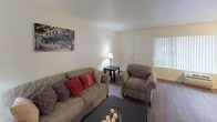 The Oak Model at 1450 Petree St in El Cajon presents a well-organized single-story layout designed for comfortable everyday living. The living room serves as the central gathering space, featuring warm wood-look flooring, neutral walls, and natural light admitted through vertical blinds. The room comfortably accommodates a sofa and armchair arrangement centered around a dark coffee table, creating an inviting and functional seating area.

The kitchen is a standout feature, finished with rich dark wood cabinetry, granite-style countertops, and recessed lighting. It is equipped with stainless steel appliances, including a dishwasher, refrigerator, and gas range, and offers ample cabinetry for storage. Light wood flooring carries through this space, complementing the warm tones of the cabinetry.

The bedroom provides a calm, restful retreat with plush carpeting, neutral walls, and sufficient space for a full-size bed and dresser with mirror. A doorway connects directly to adjacent areas, ensuring ease of movement throughout the unit. The bathroom includes a clean, tiled tub-shower combination with crisp white finishes. Overall, The Oak Model offers a practical and cohesive design suited for straightforward, comfortable living.