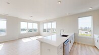 Located on the sixth floor at 100 Avenue G in Bayonne, NJ, this well-appointed 2-bedroom, 2-bathroom residence offers a bright, modern living experience. The open-concept kitchen and living area serves as the heart of the home, where a large white quartz island with an integrated sink anchors the space. The kitchen is outfitted with stainless steel appliances, including a French door refrigerator, electric range, built-in microwave, and dishwasher, complemented by a two-tone cabinet scheme of white and natural wood-grain finishes set against a clean subway tile backsplash. Light wood-tone flooring flows seamlessly throughout the unit, enhancing the airy, contemporary feel. The expansive living area is bathed in natural light from a bank of large windows offering sweeping panoramic views of the Bayonne skyline and surrounding landscape. The spacious primary bedroom enjoys its own generous window framing open outdoor views, while the second bedroom is equally well-lit. Both bathrooms are finished with light wood vanities, white quartz countertops, LED-backlit mirrors, and tiled shower surrounds. A dedicated closet provides additional storage, and a well-connected hallway ties the layout together efficiently.