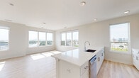 Located on the sixth floor at 100 Avenue G in Bayonne, NJ, this well-appointed 2-bedroom, 2-bathroom residence offers a bright, modern living experience. The open-concept kitchen and living area serves as the heart of the home, where a large white quartz island with an integrated sink anchors the space. The kitchen is outfitted with stainless steel appliances, including a French door refrigerator, electric range, built-in microwave, and dishwasher, complemented by a two-tone cabinet scheme of white and natural wood-grain finishes set against a clean subway tile backsplash. Light wood-tone flooring flows seamlessly throughout the unit, enhancing the airy, contemporary feel. The expansive living area is bathed in natural light from a bank of large windows offering sweeping panoramic views of the Bayonne skyline and surrounding landscape. The spacious primary bedroom enjoys its own generous window framing open outdoor views, while the second bedroom is equally well-lit. Both bathrooms are finished with light wood vanities, white quartz countertops, LED-backlit mirrors, and tiled shower surrounds. A dedicated closet provides additional storage, and a well-connected hallway ties the layout together efficiently.