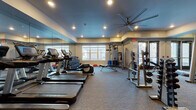 Located at 150 Country Squire Dr in Cromwell, Connecticut, this fitness center is a comprehensively equipped facility designed to support a full range of workout routines. The space features a neutral color palette with a distinctive blue-toned ceiling, recessed lighting, and a large ceiling fan that contributes to a comfortable training environment. Floor-to-ceiling mirrors line the walls, visually expanding the space and allowing users to monitor their form throughout their workouts. Large windows allow ample natural light to fill the room while providing views of the surrounding exterior. The cardio section is outfitted with multiple Life Fitness treadmills, stair climbers, and additional cardio machines, all equipped with digital display consoles. The free weights area includes a well-stocked dumbbell rack with a wide range of weights, accompanied by adjustable benches for a variety of exercises. The strength training zone features cable machines, weight racks with plate-loaded barbells, and additional resistance equipment to support both functional and traditional strength training. The flooring throughout is durable, low-pile carpet suited for gym use. A dedicated entry door provides direct access to adjacent facilities, making this fitness center a practical and well-appointed amenity.